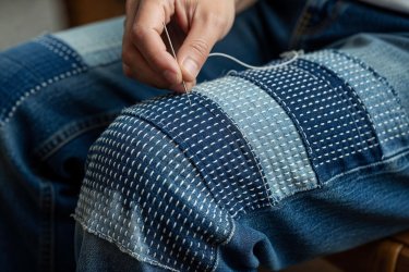 Hand holding a sashiko needle sewing white parallel stitches over indigo patched denim